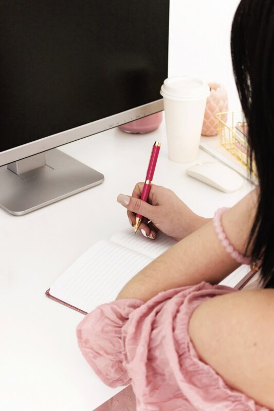 woman at desk in front of a computer writing in a notebook